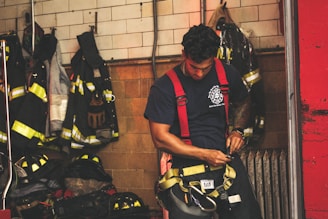 A firefighter in a navy-blue t-shirt and red suspenders is preparing his gear in a room filled with hanging protective fire jackets and helmets. The background consists of tile and brick walls, with fire equipment neatly organized.