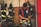 A firefighter in a navy-blue t-shirt and red suspenders is preparing his gear in a room filled with hanging protective fire jackets and helmets. The background consists of tile and brick walls, with fire equipment neatly organized.