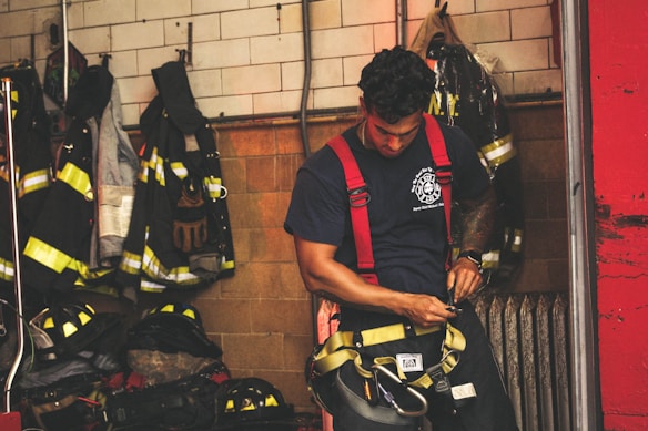 A firefighter in a navy-blue t-shirt and red suspenders is preparing his gear in a room filled with hanging protective fire jackets and helmets. The background consists of tile and brick walls, with fire equipment neatly organized.