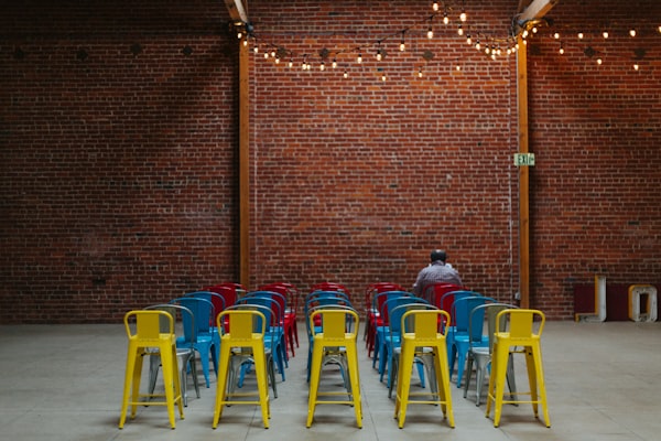 several rows of empty metal chairs with one figure sitting near the front, by a bare brick wall