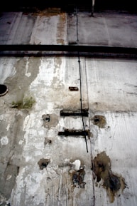 A weathered and deteriorating industrial wall features peeling paint, rust spots, and exposed metal rods. There is a black cable running vertically across the surface, and visible patches of mold or mildew contribute a sense of decay.
