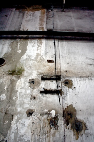 A weathered and deteriorating industrial wall features peeling paint, rust spots, and exposed metal rods. There is a black cable running vertically across the surface, and visible patches of mold or mildew contribute a sense of decay.
