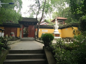 A serene stone courtyard featuring traditional Chinese architecture is surrounded by lush green trees. In the foreground, stone steps lead up to a doorway adorned with red lanterns and Chinese inscriptions. A white stone statue stands to the right, nestled among the greenery and yellow building walls. A small, tiled-roof pavilion is visible in the background.