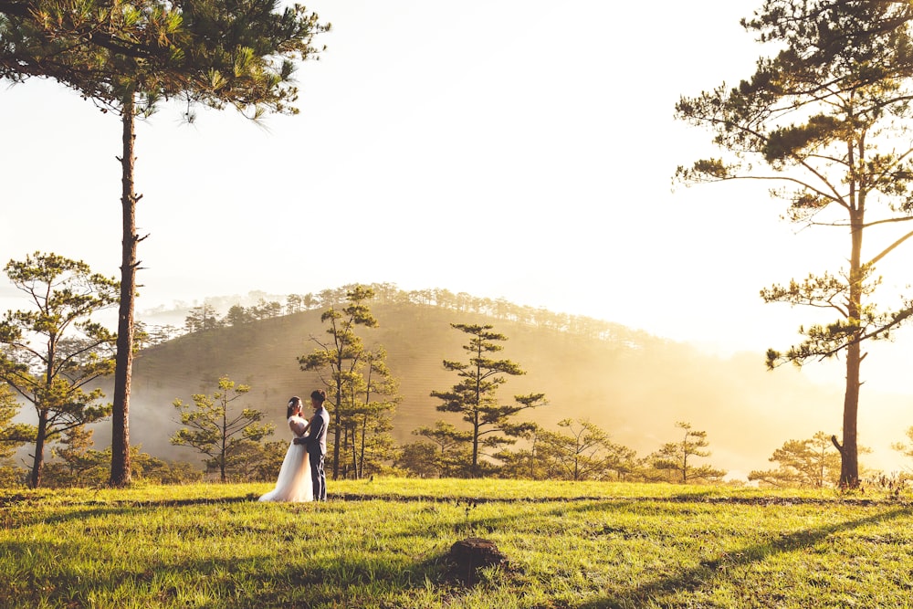 Le marié et la mariée debout sur un champ d’herbe verte tout en se câlinant et avec la montagne et les arbres au loin pendant la journée