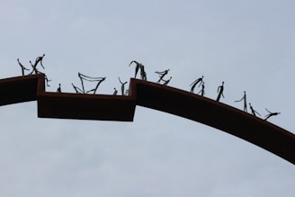 A group of abstract figures walks along a curved, elevated pathway. The figures are simplified, resembling stick figures, and appear to be sculpted or painted in a dark color against a light sky background. The pathway is broken into sections with gaps between them, suggesting themes of separation or journey.
