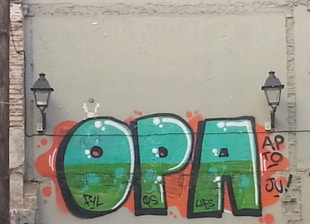 Graffiti artwork featuring large, colorful letters spelling 'OPA' on a textured wall. The letters have a vibrant green color with light blue highlights and are outlined in black. Each letter is surrounded by red-orange splashes, giving a dynamic effect. Two black wall-mounted lamps are positioned on either side of the graffiti.