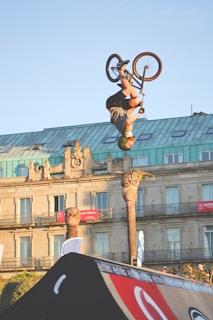 A person is performing an impressive backflip on a BMX bike in mid-air against a backdrop of an old architectural building with ornate designs. Palm trees are seen beside the structure, and the scene is lit by soft daylight, highlighting the green roof and beige facade of the building.