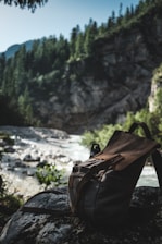 A rustic backpack rests on a rock beside a flowing river, set against a backdrop of lush, evergreen forest and rugged mountains under a clear sky. The scene is shadowy and serene, suggesting an untouched and natural environment.