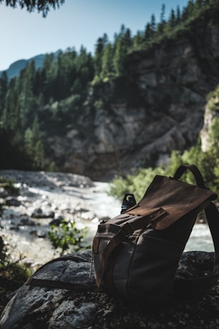 A rustic backpack rests on a rock beside a flowing river, set against a backdrop of lush, evergreen forest and rugged mountains under a clear sky. The scene is shadowy and serene, suggesting an untouched and natural environment.