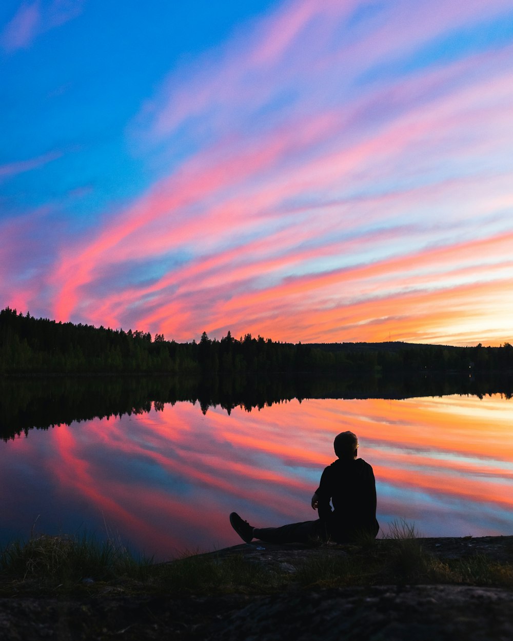 silhouette of man sitting on water's edge facing mountain during golden hour