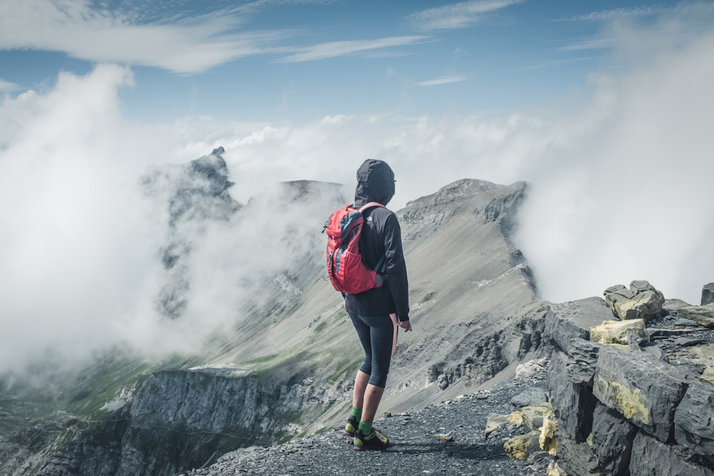 man wearing black hooded jacket and backpack standing on rocks facing clouds