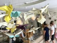 Origami birds in various colors, such as yellow, blue, white, and purple, hang from the ceiling. The background shows an indoor cafe or shop setting with blurred figures of people standing at a counter.