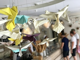 Origami birds in various colors, such as yellow, blue, white, and purple, hang from the ceiling. The background shows an indoor cafe or shop setting with blurred figures of people standing at a counter.