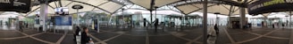 A wide-angle view of a modern transportation hub with a covered walkway, display boards, and seating areas. Glass walls offer a view of the surrounding cityscape. Several people are seated or walking, and signs and advertisements are visible.