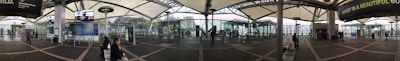 A wide-angle view of a modern transportation hub with a covered walkway, display boards, and seating areas. Glass walls offer a view of the surrounding cityscape. Several people are seated or walking, and signs and advertisements are visible.