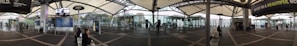 A wide-angle view of a modern transportation hub with a covered walkway, display boards, and seating areas. Glass walls offer a view of the surrounding cityscape. Several people are seated or walking, and signs and advertisements are visible.
