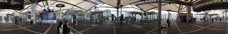 A wide-angle view of a modern transportation hub with a covered walkway, display boards, and seating areas. Glass walls offer a view of the surrounding cityscape. Several people are seated or walking, and signs and advertisements are visible.