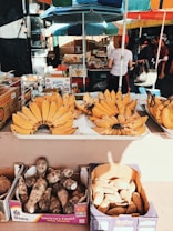 A market stall featuring bunches of ripe bananas arranged in trays, surrounded by boxes filled with root vegetables like yams. The stand is set under colorful umbrellas, and people in casual clothing are seen shopping in the background. The atmosphere is vibrant and bustling.