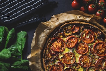 A rustic quiche or tart topped with sliced tomatoes is placed on parchment paper. Surrounding the dish, there are fresh tomatoes on the vine and a bunch of spinach leaves. A black and white striped cloth adds to the warm, homely atmosphere of the image.