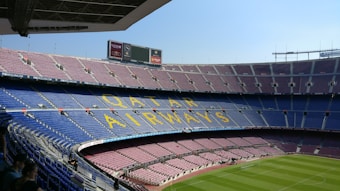 A large, empty football stadium with multiple tiers of seating. The seats are mostly blue and maroon, with 'Qatar Airways' written in large yellow letters across the seats on one tier. The field is well-maintained with bright green grass. There are electronic advertising boards and a few people sitting in the lower stands.