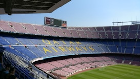 A large, empty football stadium with multiple tiers of seating. The seats are mostly blue and maroon, with 'Qatar Airways' written in large yellow letters across the seats on one tier. The field is well-maintained with bright green grass. There are electronic advertising boards and a few people sitting in the lower stands.