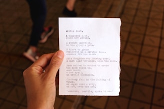 A person's hand is holding a piece of paper with typed text, featuring a poem. In the background, the blurred image of a person walking and a tiled floor can be seen.