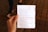 A person's hand is holding a piece of paper with typed text, featuring a poem. In the background, the blurred image of a person walking and a tiled floor can be seen.