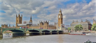 A panoramic view of the iconic Big Ben and the Houses of Parliament in London. The scene includes the Westminster Bridge spanning the River Thames, with double-decker buses and other vehicles crossing. The buildings exhibit Gothic architecture with intricate details. The sky is partly cloudy, casting a soft light over the river and the structures.