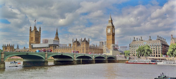 A panoramic view of the iconic Big Ben and the Houses of Parliament in London. The scene includes the Westminster Bridge spanning the River Thames, with double-decker buses and other vehicles crossing. The buildings exhibit Gothic architecture with intricate details. The sky is partly cloudy, casting a soft light over the river and the structures.
