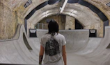 A person stands in an indoor skate park with concrete ramps and curved surfaces. The park is located in a tunnel with brick walls and metal signage, illuminated by fluorescent lights. The person is wearing a white t-shirt with a graphic design and has tattoos on their arms.