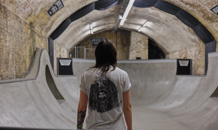 A person stands in an indoor skate park with concrete ramps and curved surfaces. The park is located in a tunnel with brick walls and metal signage, illuminated by fluorescent lights. The person is wearing a white t-shirt with a graphic design and has tattoos on their arms.