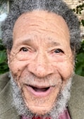 An older man with a curly gray beard and hair is smiling broadly. His face is lined with wrinkles, and he has a joyful expression. The background consists of greenery, suggesting an outdoor setting.