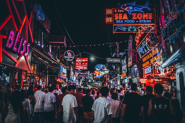 A bustling street scene at night, crowded with people walking beneath a multitude of colorful neon signs advertising various businesses such as restaurants and clubs. The vivid signs feature text and images, contributing to a lively and vibrant atmosphere.