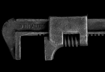 A detailed close-up image of a metal adjustable wrench with a textured surface and visible screw mechanism. The wrench is positioned horizontally against a solid black background, which highlights its rugged and industrial appearance.