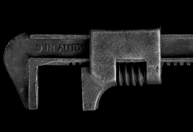 A detailed close-up image of a metal adjustable wrench with a textured surface and visible screw mechanism. The wrench is positioned horizontally against a solid black background, which highlights its rugged and industrial appearance.