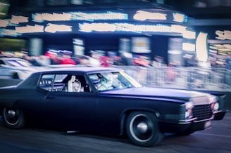 A classic black car is in motion on a busy street, surrounded by blurred neon lights and a crowd of people. The scene conveys a sense of urban speed and excitement, with a focus on the sleek vehicle. The car's headlights and the surrounding lights create a vivid atmosphere.