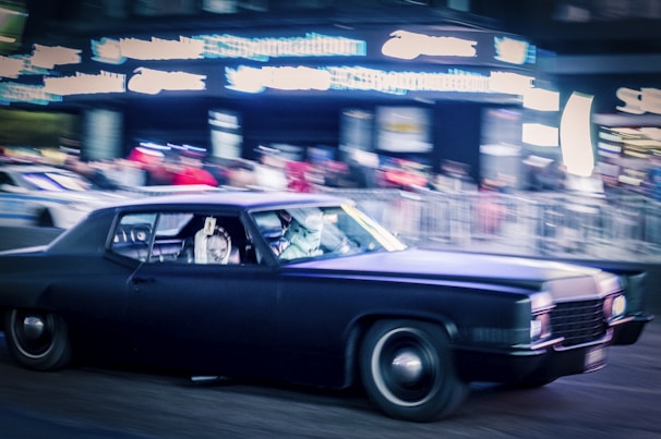A classic black car is in motion on a busy street, surrounded by blurred neon lights and a crowd of people. The scene conveys a sense of urban speed and excitement, with a focus on the sleek vehicle. The car's headlights and the surrounding lights create a vivid atmosphere.