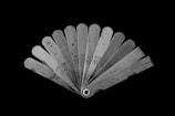 A fan-like arrangement of metal feeler gauges spread out, each labeled with different measurements in millimeters. The gauges are metallic with a slightly reflective surface, showing signs of wear. The background is stark black, highlighting the tools.