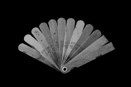 A fan-like arrangement of metal feeler gauges spread out, each labeled with different measurements in millimeters. The gauges are metallic with a slightly reflective surface, showing signs of wear. The background is stark black, highlighting the tools.