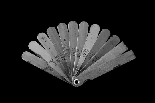 A fan-like arrangement of metal feeler gauges spread out, each labeled with different measurements in millimeters. The gauges are metallic with a slightly reflective surface, showing signs of wear. The background is stark black, highlighting the tools.