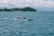 A small fishing boat is navigating through a clear, expansive ocean with gentle waves. In the background, a lush, green island is visible under a partly cloudy sky. The horizon is faintly marked by distant mountains and more islands, creating a serene, picturesque seascape.