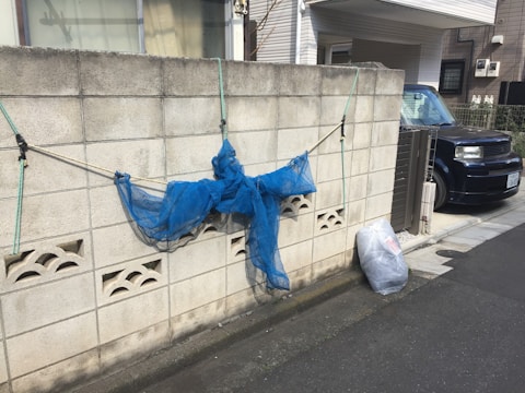 A gray concrete wall with a blue net draped over it, held in place by green and black ropes. Below the net, a white plastic bag is placed on the ground. A dark-colored car is parked on the right side, next to a driveway and a house.