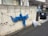 A gray concrete wall with a blue net draped over it, held in place by green and black ropes. Below the net, a white plastic bag is placed on the ground. A dark-colored car is parked on the right side, next to a driveway and a house.