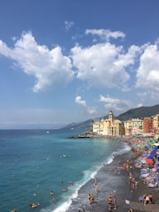 A picturesque coastal town with vibrant, colorful buildings lining a beach. Numerous people are enjoying the sandy shore and swimming in the turquoise sea. The sky is bright with large, fluffy clouds, and the mountains are visible in the background.