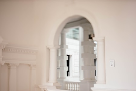 An elegant interior featuring white classical architecture with arches, columns, and decorative moldings. Soft lighting casts subtle shadows, enhancing the refined and minimalistic aesthetic.
