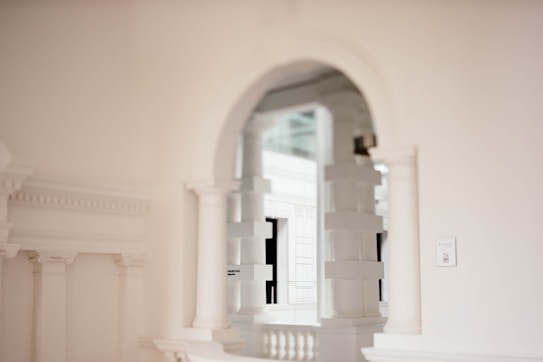 An elegant interior featuring white classical architecture with arches, columns, and decorative moldings. Soft lighting casts subtle shadows, enhancing the refined and minimalistic aesthetic.