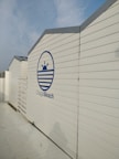 A series of white, wooden beach cabins with a blue logo that includes a stylized sun and waves, and the text 'Chagall Beach'. The cabins are aligned in a row under a partly cloudy blue sky.