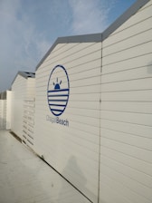 A series of white, wooden beach cabins with a blue logo that includes a stylized sun and waves, and the text 'Chagall Beach'. The cabins are aligned in a row under a partly cloudy blue sky.