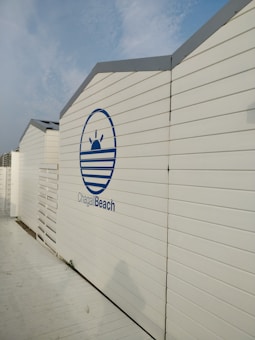 A series of white, wooden beach cabins with a blue logo that includes a stylized sun and waves, and the text 'Chagall Beach'. The cabins are aligned in a row under a partly cloudy blue sky.