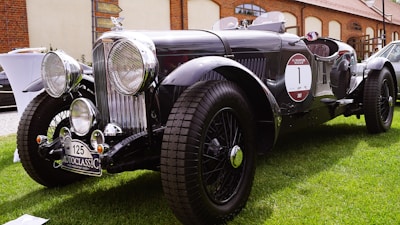 A vintage black classic car with prominent circular headlights and chrome details is parked on a lush green lawn. It features a distinct front grille, large tires, and a 'MotoClassic' license plate. The surroundings include a brick building and various other vehicles.